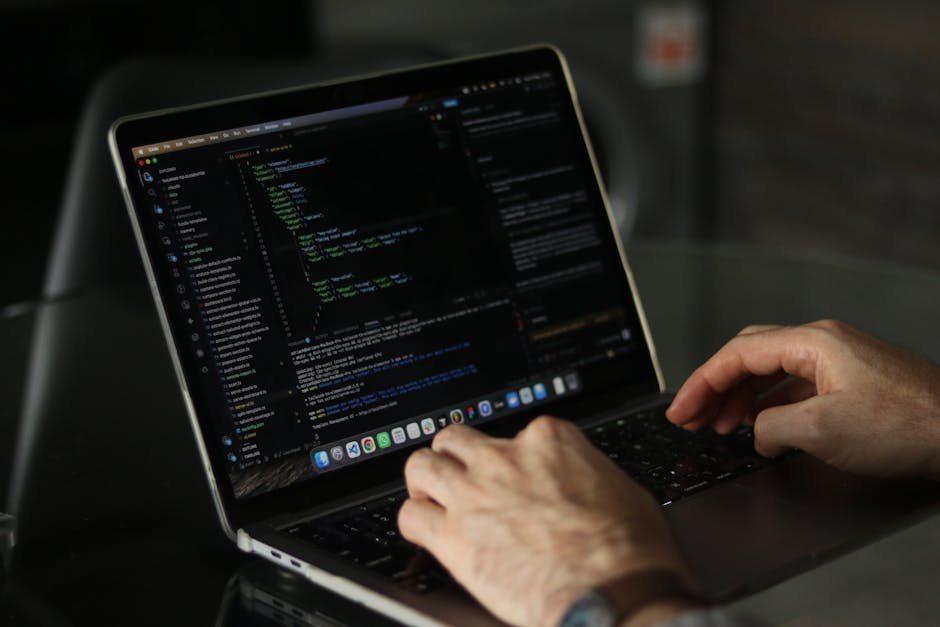 Close-up of a person's hands working on a laptop with a dark-themed code editor displayed on the screen, showing lines of programming code with colored syntax highlighting. The laptop is placed on a d